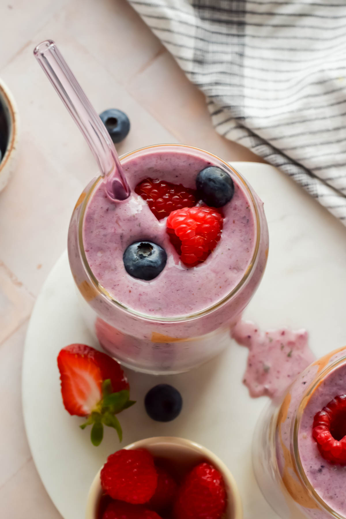 berry smoothie in clear glass with glass straw topped with fresh raspberries and blueberries.