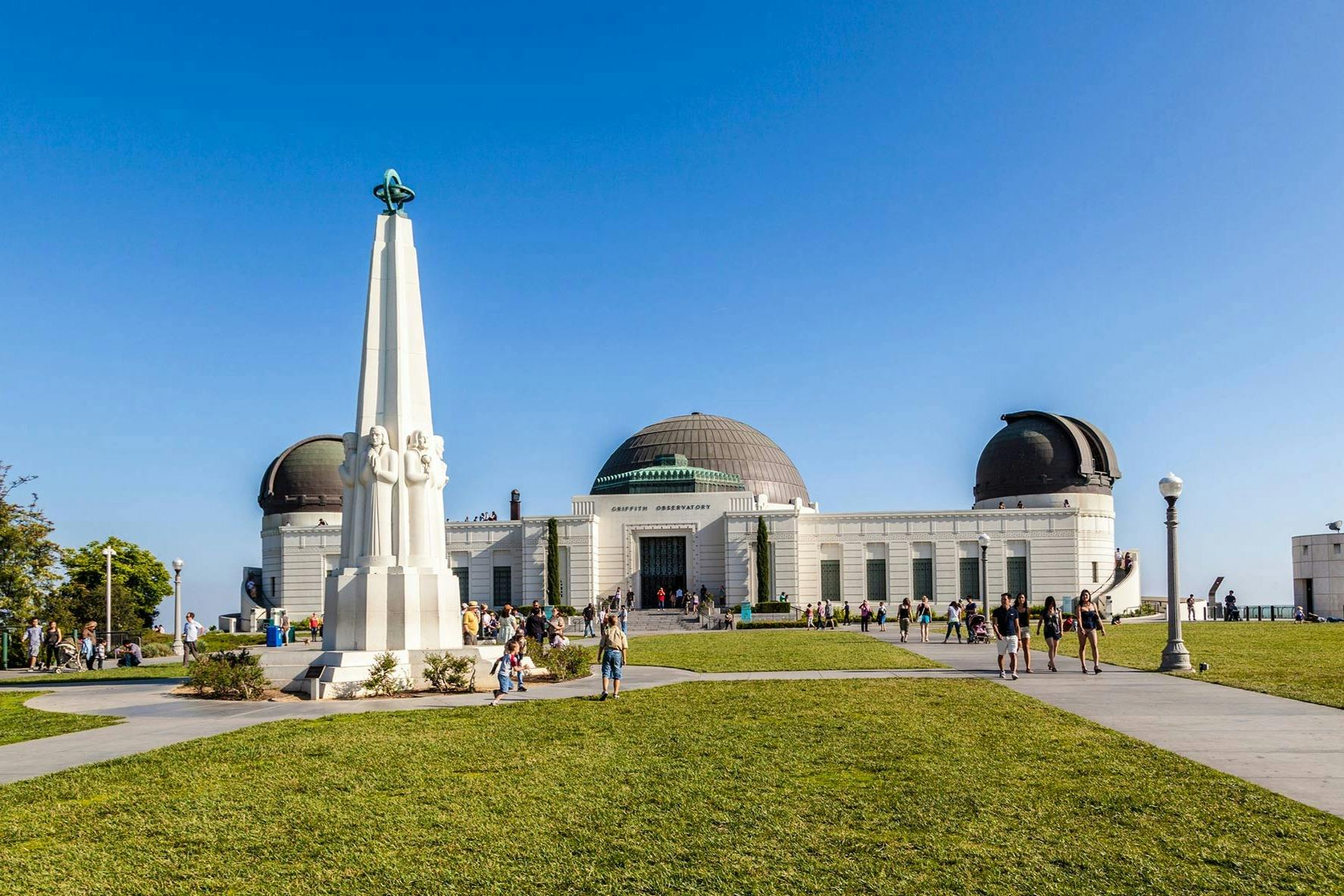A large white observatory with domes, fronted by a tall obelisk and grassy area filled with people on a clear day.