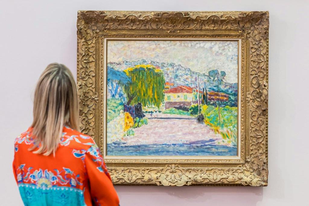 A woman in a colorful shirt views an ornate-framed painting of a serene landscape, featuring a house, trees, and a pathway.