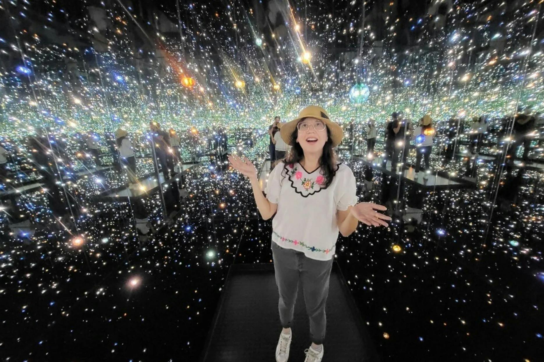 A woman stands in a room filled with mirrored walls and twinkling lights, wearing a hat, glasses, floral top, gray pants, and sneakers.
