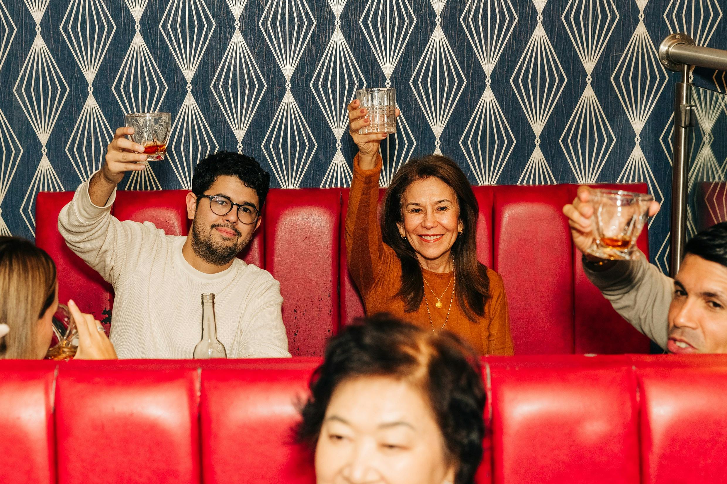 Three people sit in a booth with red seats, smiling and raising glasses. A patterned wallpaper is visible in the background.