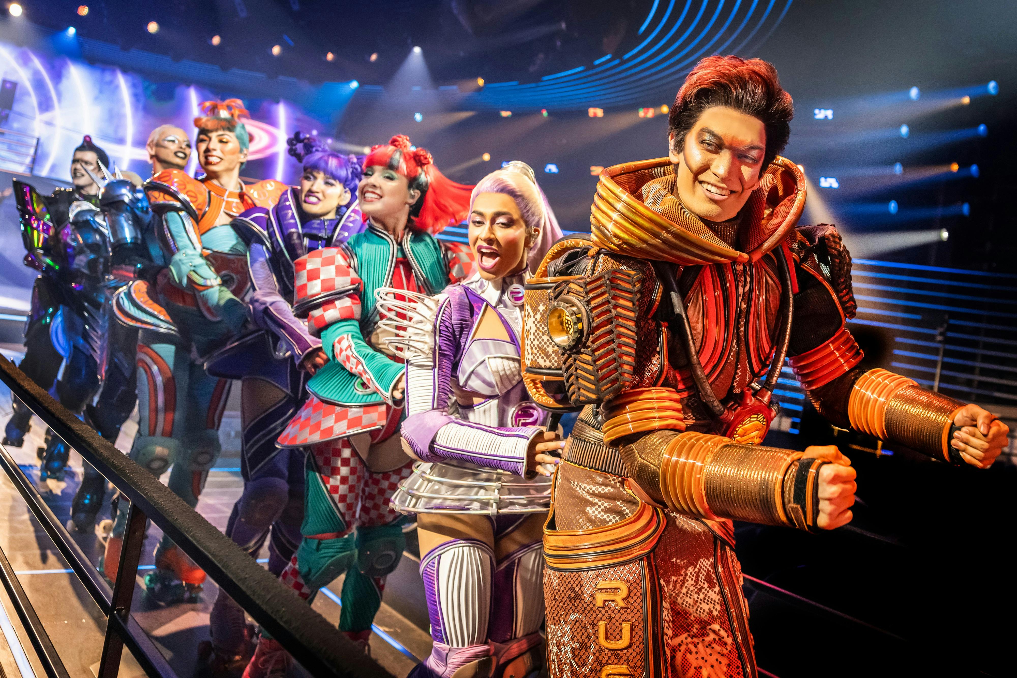 People in elaborate, futuristic costumes perform on a brightly lit stage with colorful spotlights.