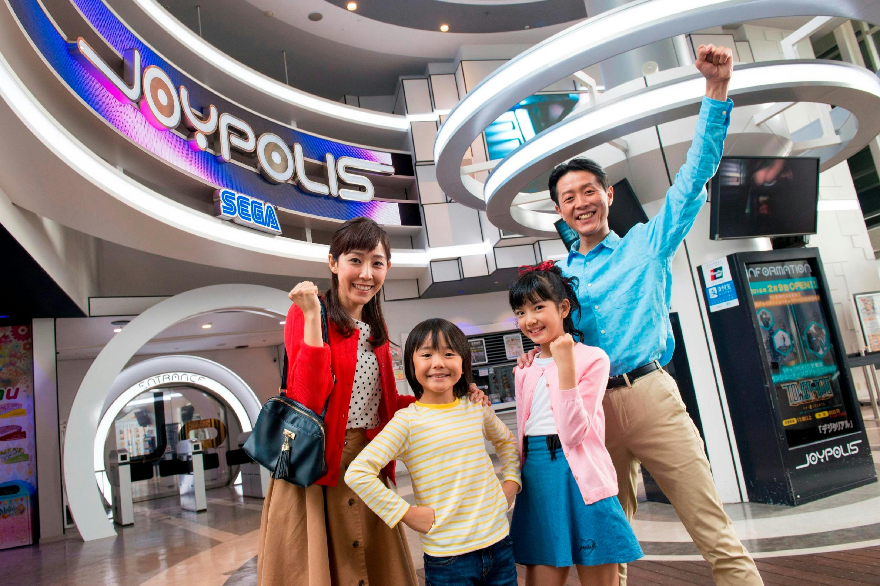A smiling family of four poses with raised fists in front of a Sega Joypolis arcade entrance.