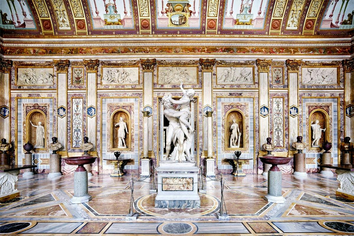 A richly decorated interior with ornate walls, statues in alcoves, and a central marble sculpture surrounded by decorative elements.