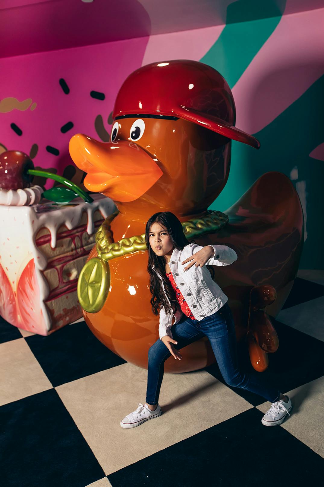 A girl posing playfully in front of a large duck statue wearing a cap, with a giant cake prop in the background.