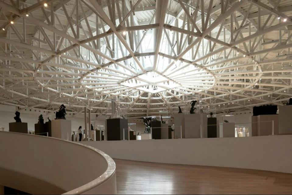 Modern art gallery with a spacious interior, featuring high, geometric ceilings and various sculptures displayed on pedestals.