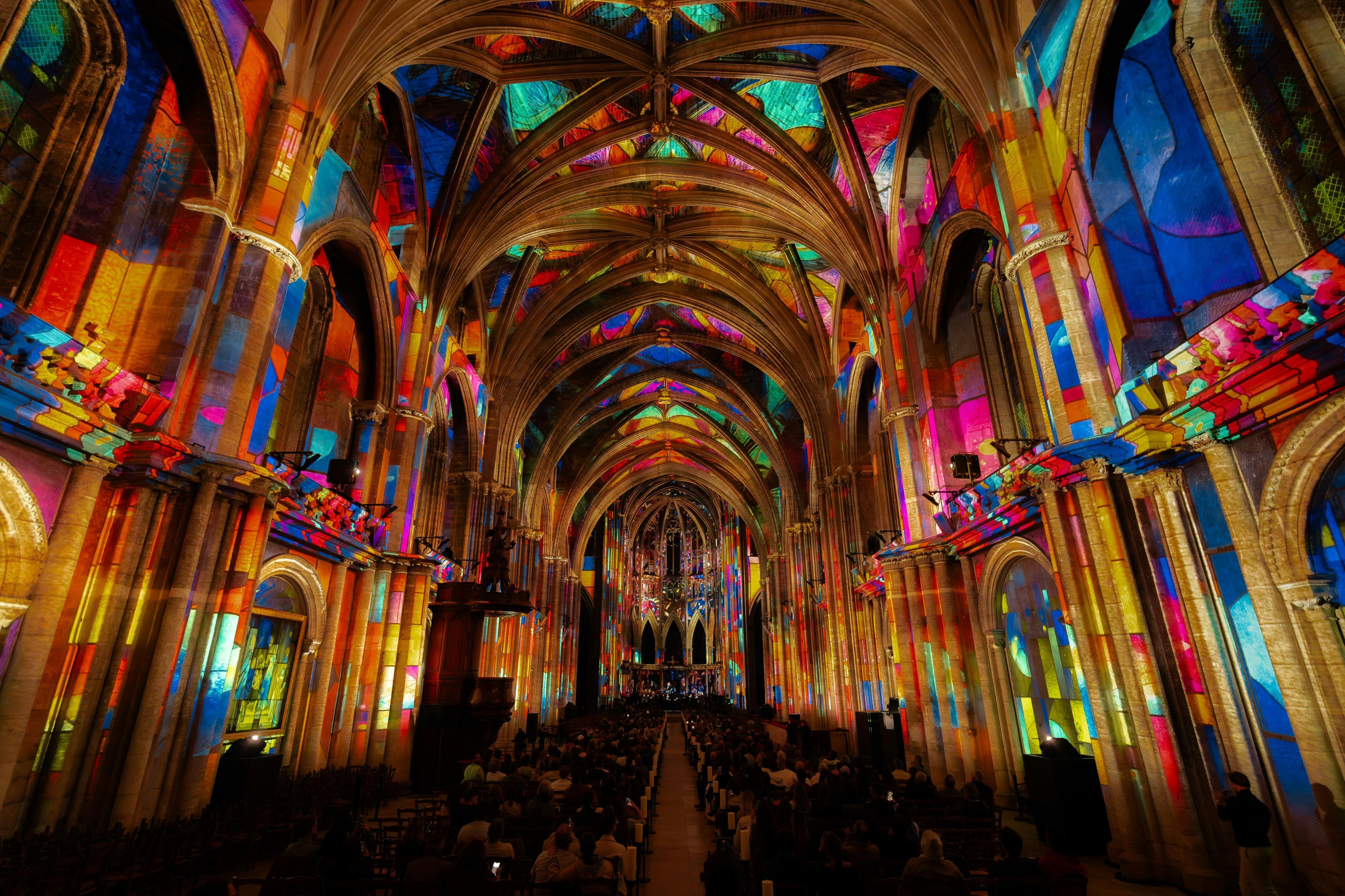 A grand cathedral interior with colorful light projections on its arches and walls, filled with seated attendees.