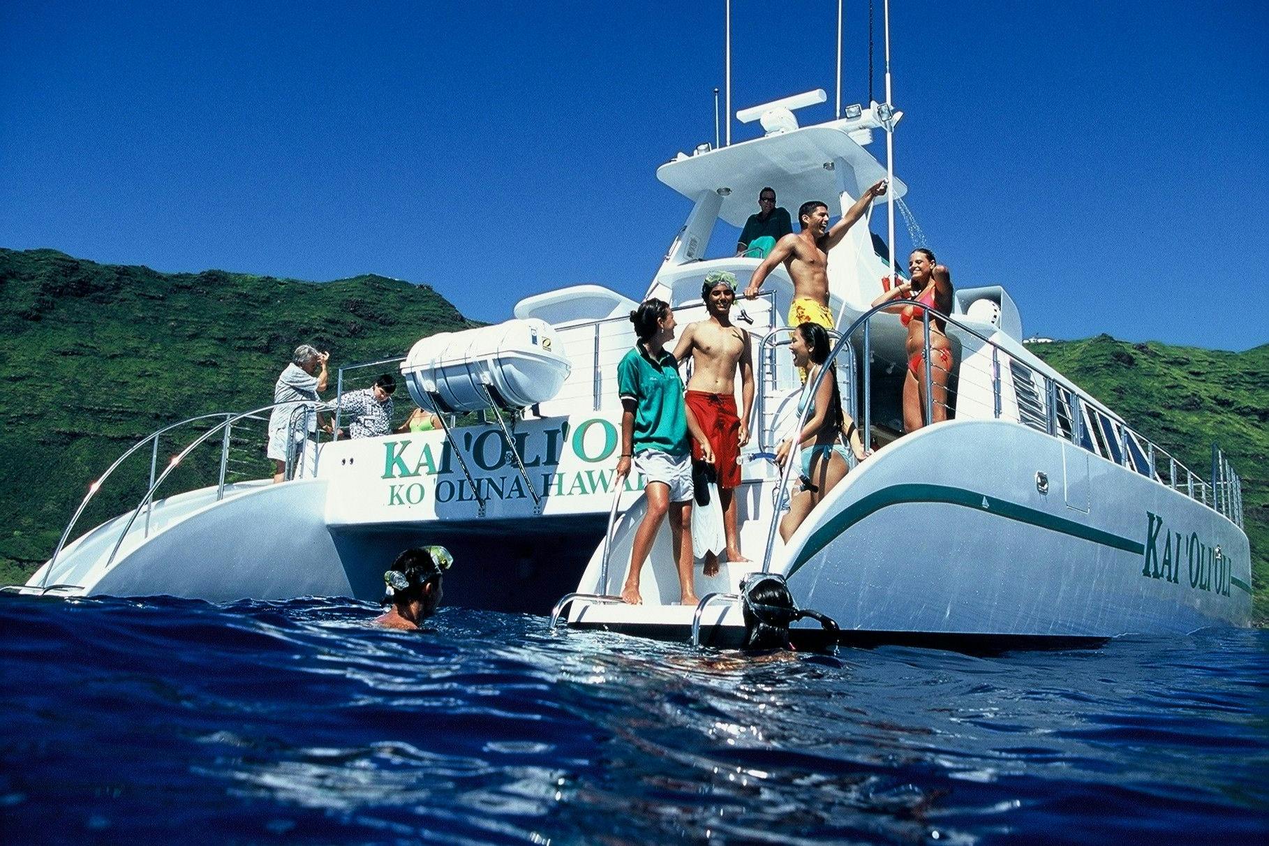 People in swimwear on a white catamaran with "Ko Olina, Hawaii" written on it, next to others snorkeling in blue water.