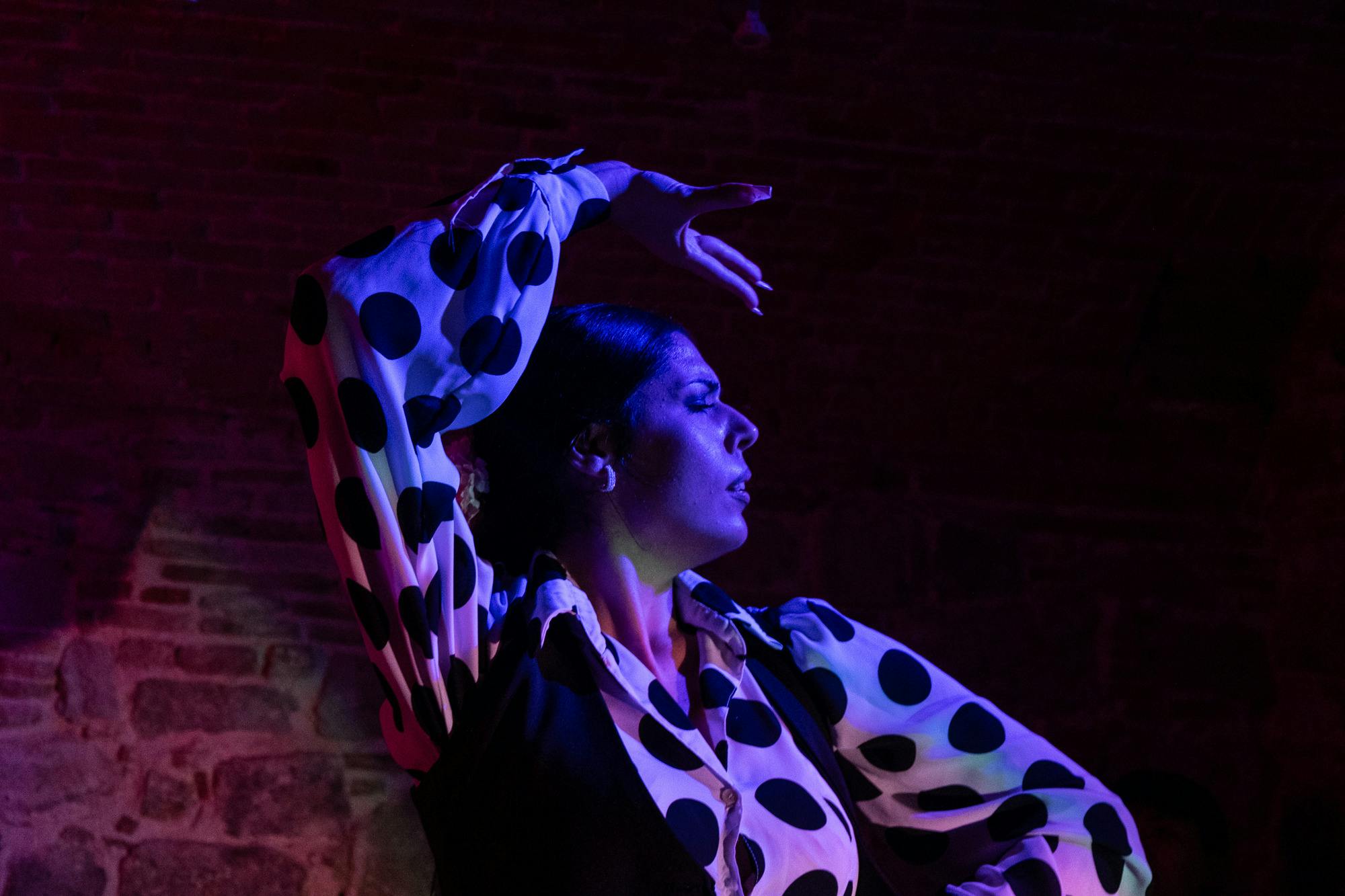 A person in a polka dot blouse raises an arm in a dance pose under dramatic lighting with a dark brick background.