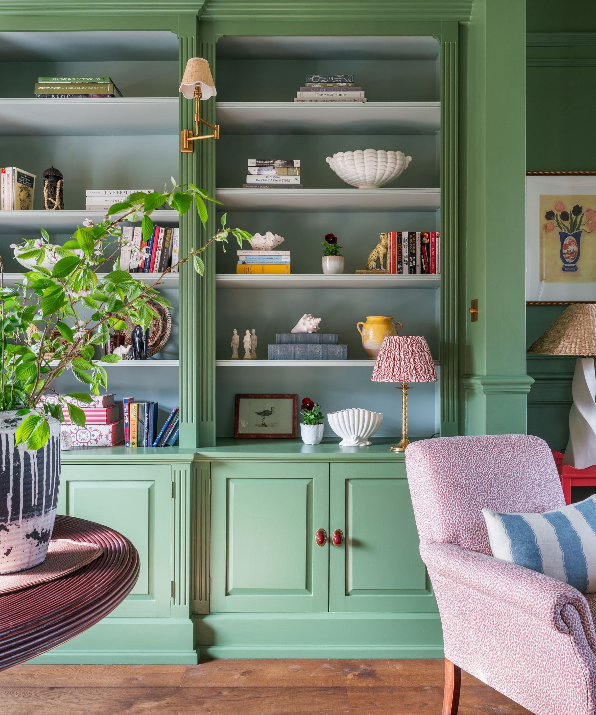 Living room with a built-in bookcase painted in Farrow & Ball's Calke green, decorated with books, ceramics, and trinkets, a pink armchair with a striped blue cushion, and a dark wood round table