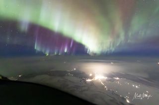 Cortinas de auroras verdes llenan el cielo, con nubes rotas debajo y resplandores distantes de las luces de la ciudad.