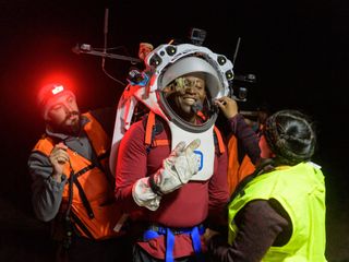 Un astronauta en el centro de la imagen lleva un casco y un traje espacial simulados. Dos ingenieros a cada lado ajustan su casco.