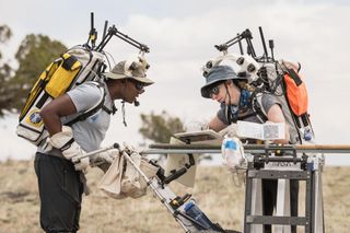 Dos astronautas inclinados sobre una mesa en un campo, mientras llevan mochilas con antenas en la parte superior.