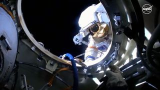 un cosmonauta con un traje espacial blanco durante una caminata espacial, visto a través de una parte circular metálica de la estación espacial internacional