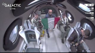 Tres personas en un avión espacial. Una de ellas sostiene una bandera italiana.