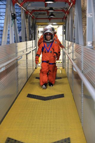 Los astronautas naranjas caminan en una línea por un pórtico amarillo.