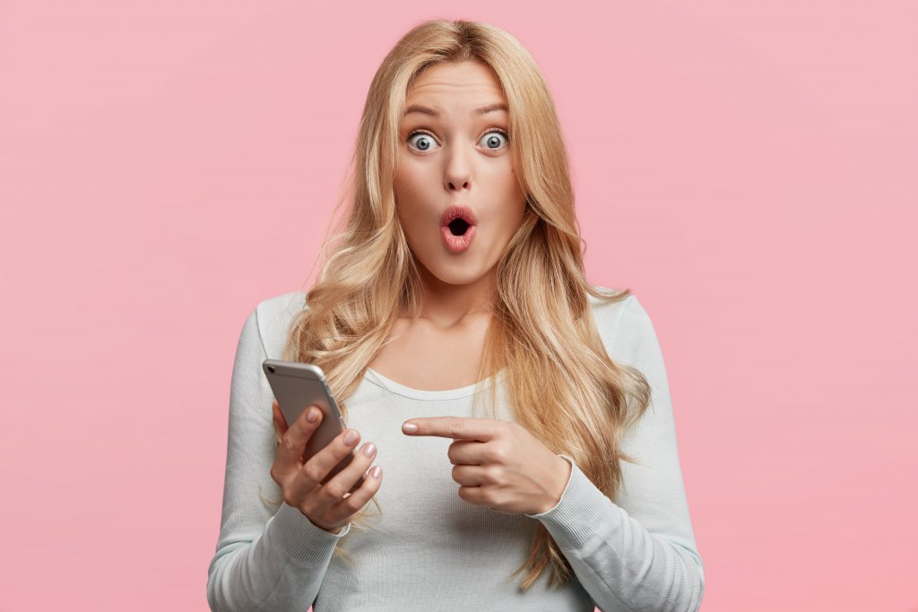 Blonde young female with pleasant appearance looks with terrified expression in smart phone, reads shocking news on webpage, isolated over pink background. Woman indicates at digital telephone