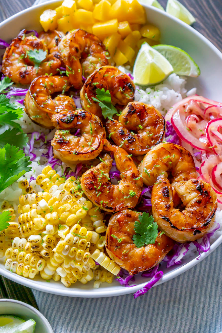 Shrimp Taco Bowl on rice with pickled onions, limes, mango, cilantro, and purple cabbage in a white bowl.