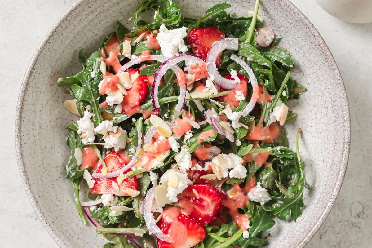 A finished strawberry arugula salad in a bowl topped with goat cheese, red onion, and fresh strawberries. 