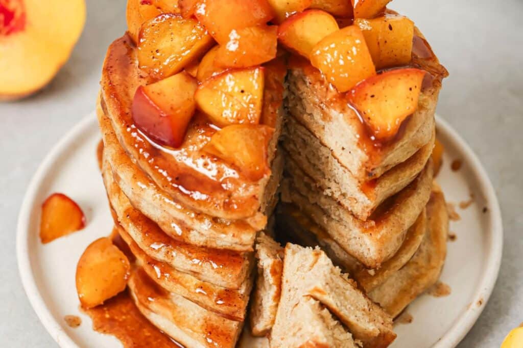 A stack of pancakes with a big portion taken out. Juicy peaches are resting on top. 