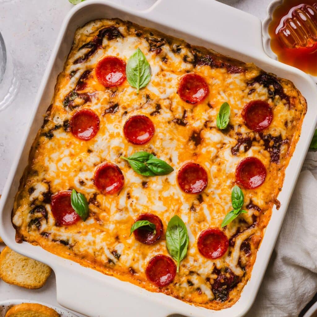 Pizza dip in a square baking dish topped with mini pepperoni, fresh basil, and a drizzle of hot honey.