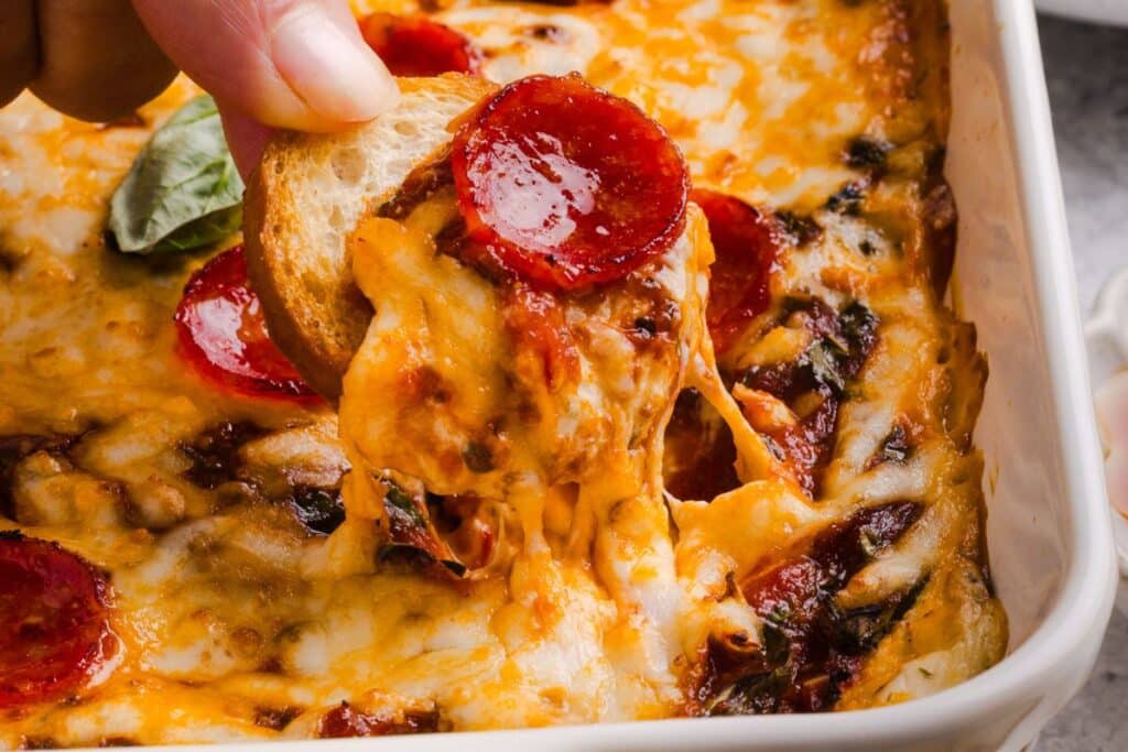 Scooping pizza dip out of a dish with a crostini. 