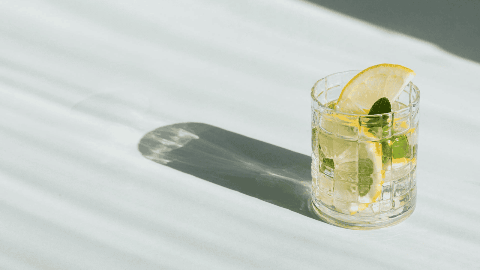 A glass of water with lemon slices and mint leaves is placed on a light surface. The glass casts a shadow on the surface. The light source creates striped patterns on the background.