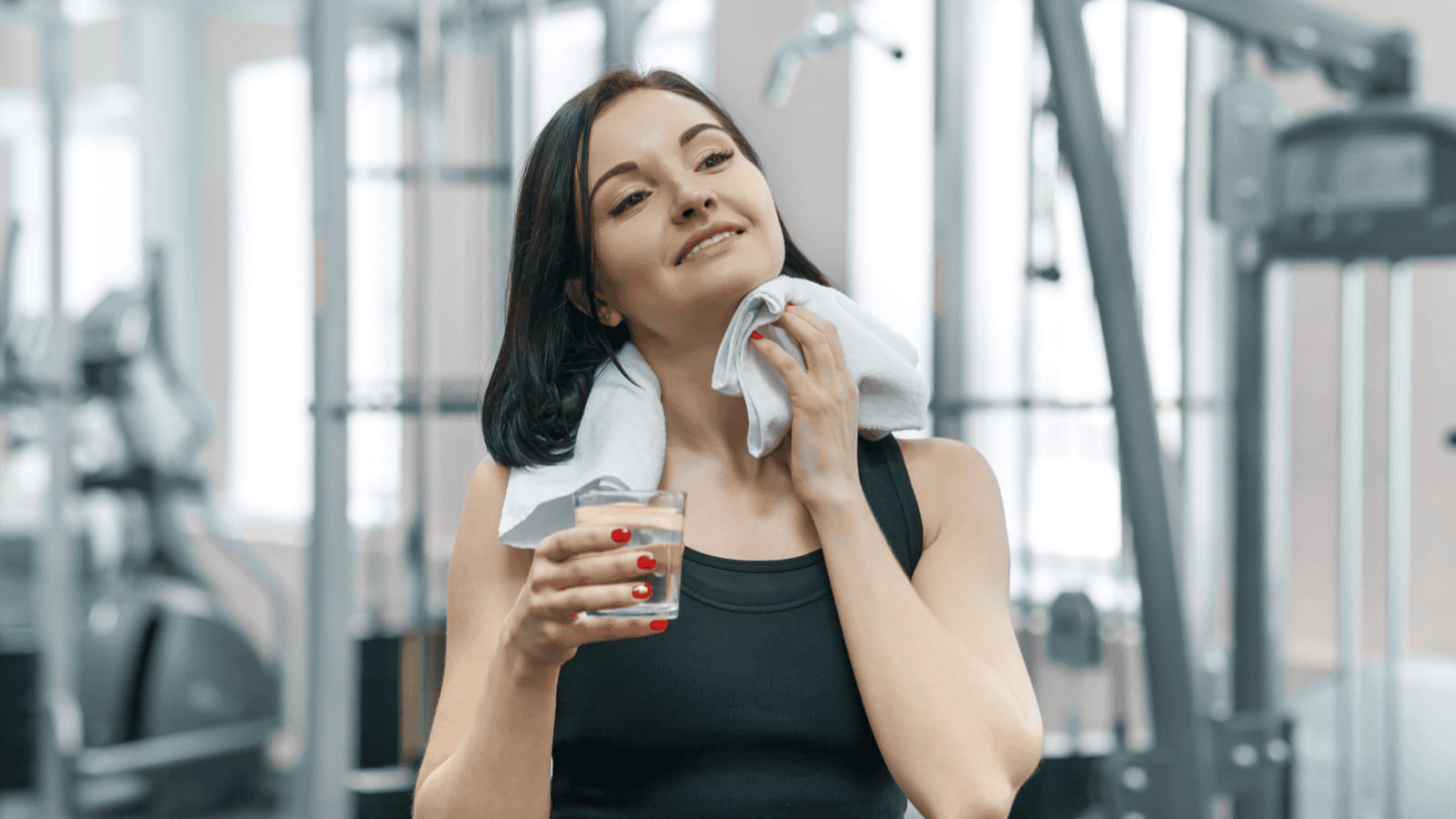 A woman with dark hair is in a gym, wearing a black tank top. She is holding a glass of a light-colored drink in one hand and a white towel in the other, which she is using to wipe her neck. Gym equipment is visible in the background.