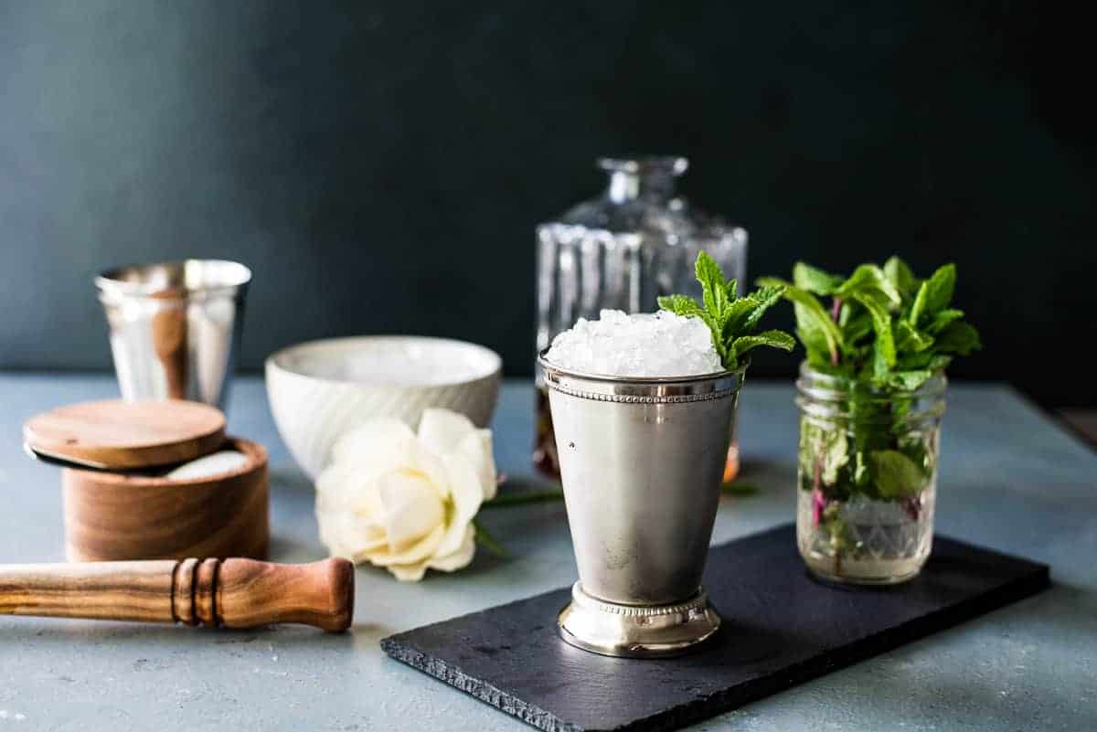 A julep cup filled with crushed ice and mint, accompanied by bartending tools, a cocktail shaker, and a fresh mint sprig in a jar, arranged on a slate board against a dark backdrop.