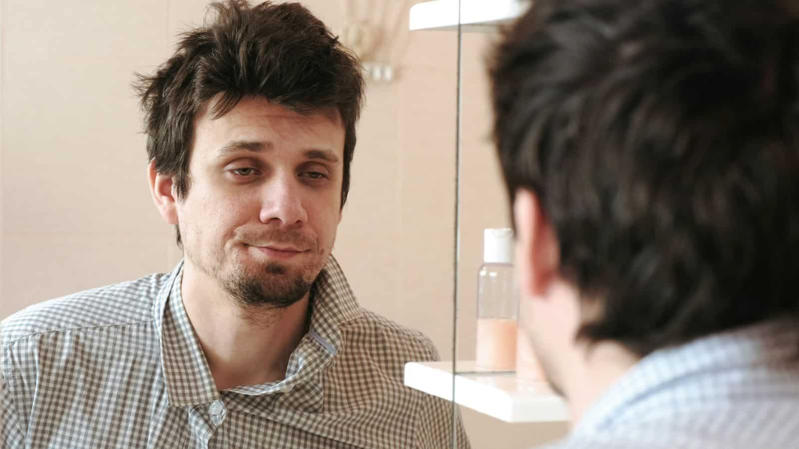 Tired and disheveled-looking man looking at his reflection in the mirror