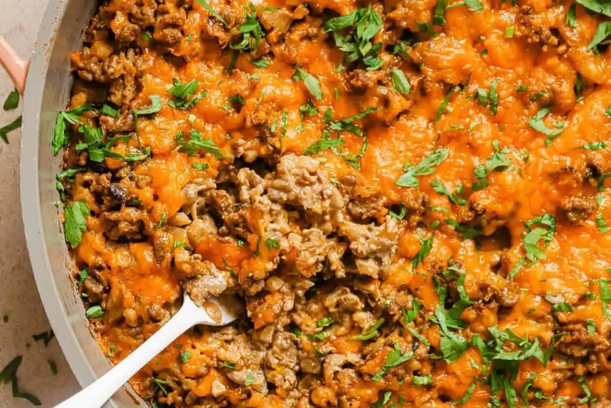 A skillet filled with a cheesy ground meat dish, topped with chopped green herbs. A spoon is partially submerged in the mixture.