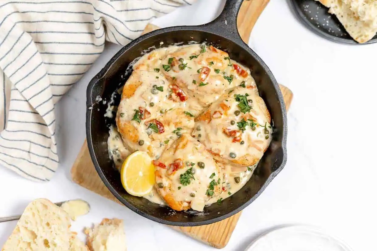 A skillet dish with creamy chicken breasts garnished with parsley, capers, and sun-dried tomatoes. A slice of lemon and some sliced bread are visible beside the pan.