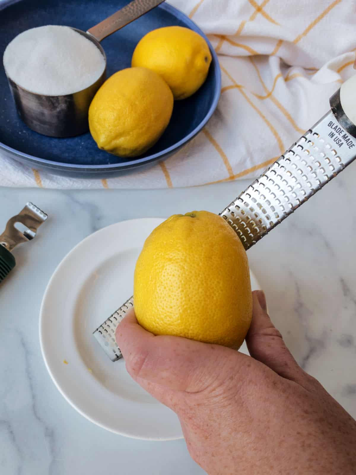 A hand grating a lemon over a white plate. Nearby are two lemons on a blue plate, a measuring cup with sugar, and a white and yellow checkered towel.