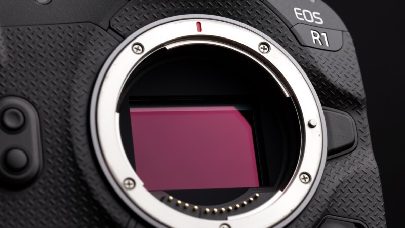 Close-up of a camera's open lens mount, showing the sensor inside. The camera body has a textured black surface with an