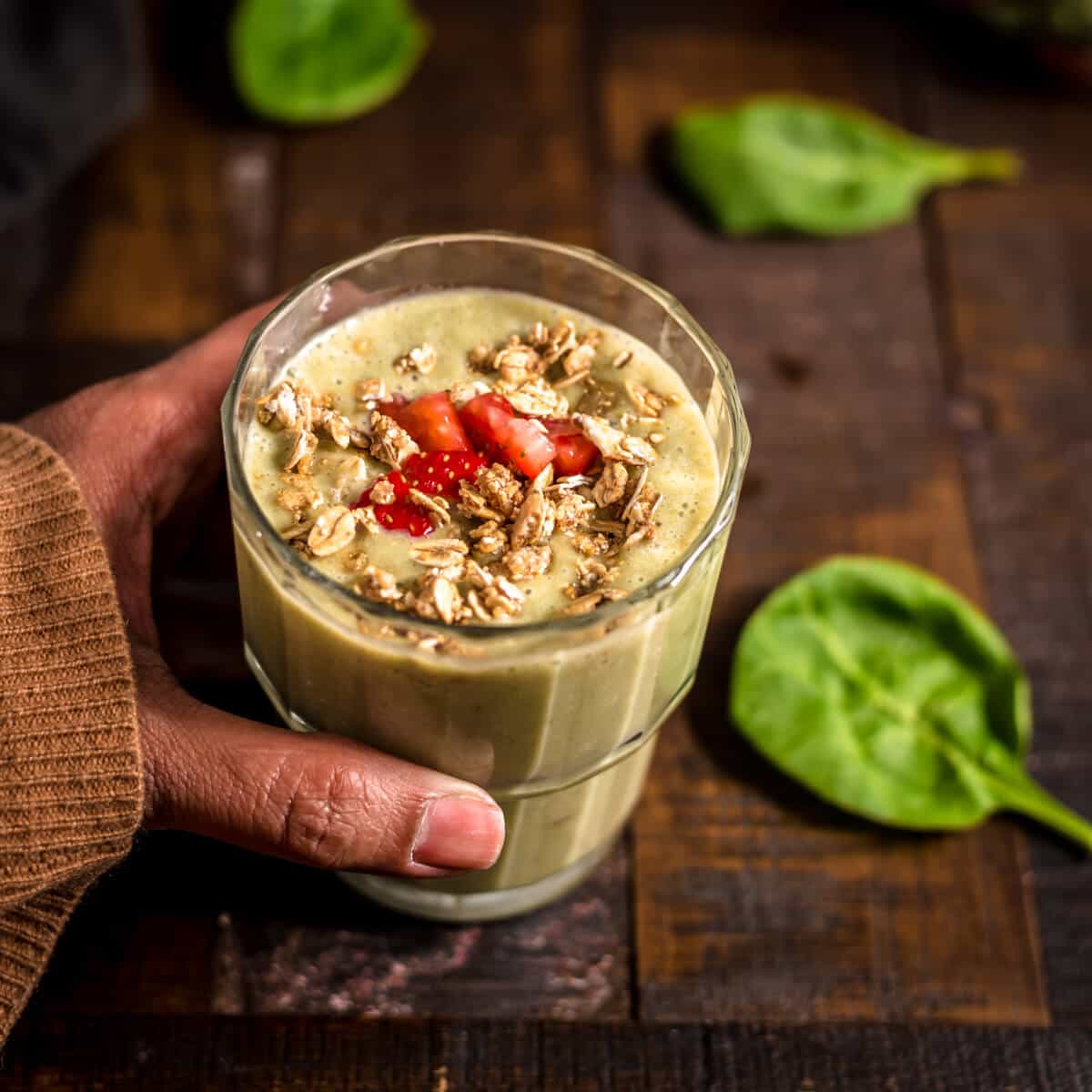 A hand holding a glass of strawberry banana spinach smoothie garnished with strawberry and granolas