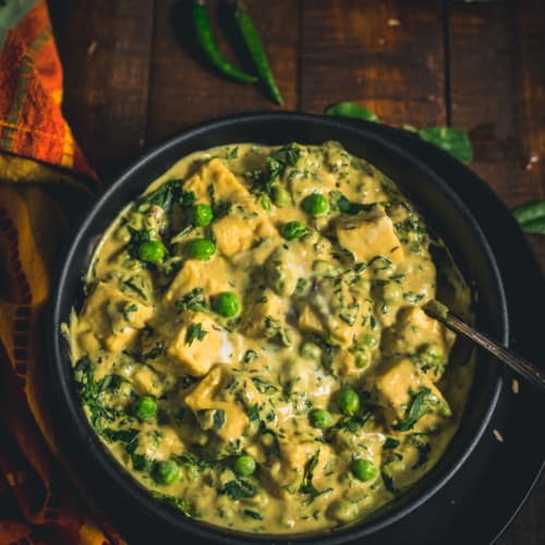 A black bowl of Methi Malai Mutter Paneer on a wooden table with green chilies and a side of naan.