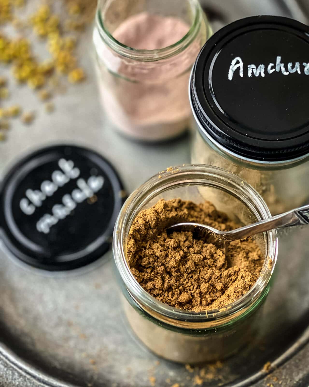 A glass bottle with chaat masala and a spoon in it. On the side is another bottle with Amchur labeled on the black cap