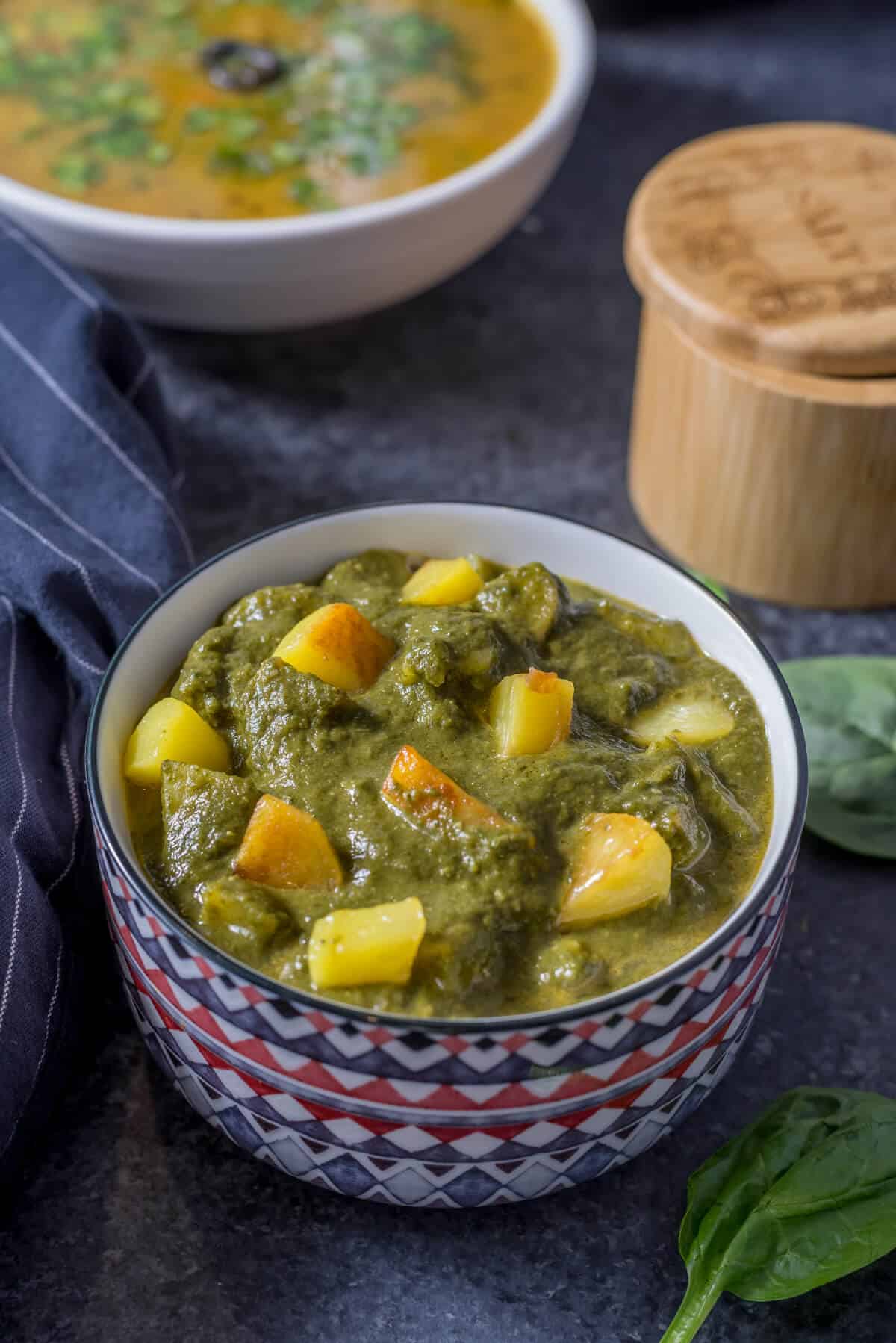 A red and white bowl with aloo palak topped with potatoes on blue counter with spinach on the side.