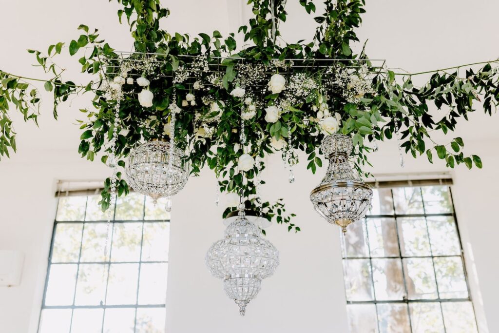 Crystal chandeliers hang in a bright, white venue surrounded by greenery