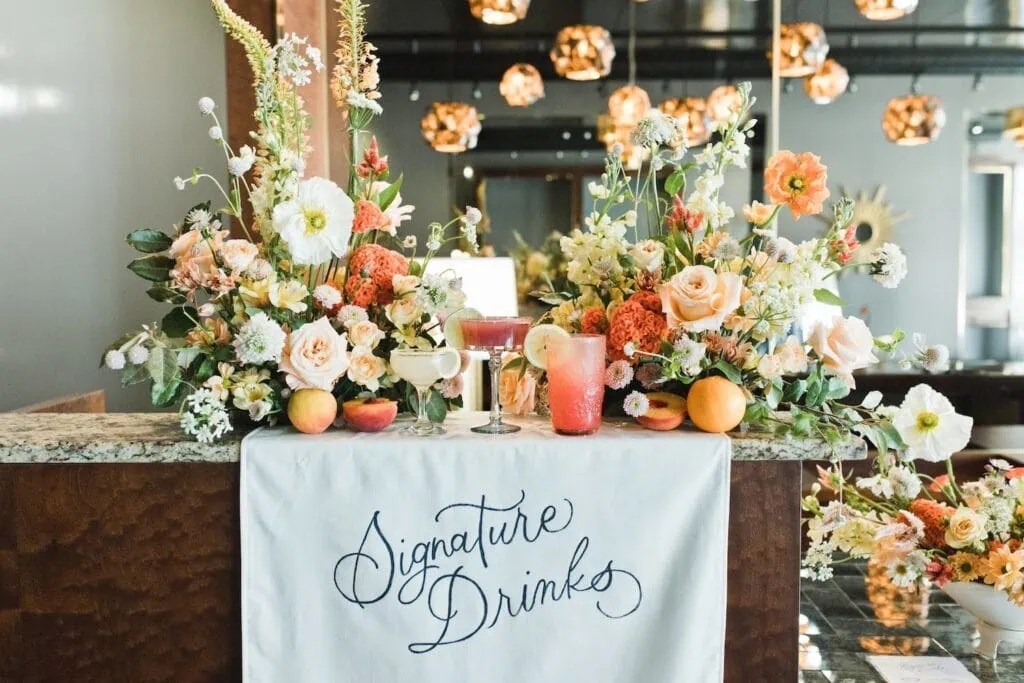 Pink, peach and white flowers decorate a bar displaying signature drinks in the same colors at a reception