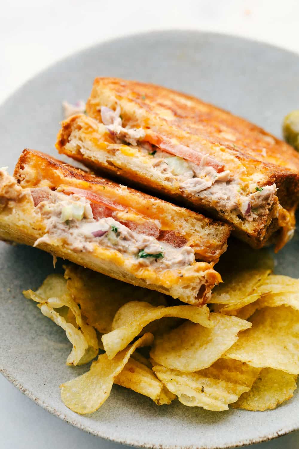 Tuna melts are the ultimate sandwich with a flavorful fish mixture, melty cheddar cheese, and freshly sliced tomatoes all melted together in a skillet on crispy, golden bread. So easy to make and delicious, this sandwich will literally melt in your mouth! Tuna melt sandwich sitting up on the plate for you to see the layers melted together with a side of chips.