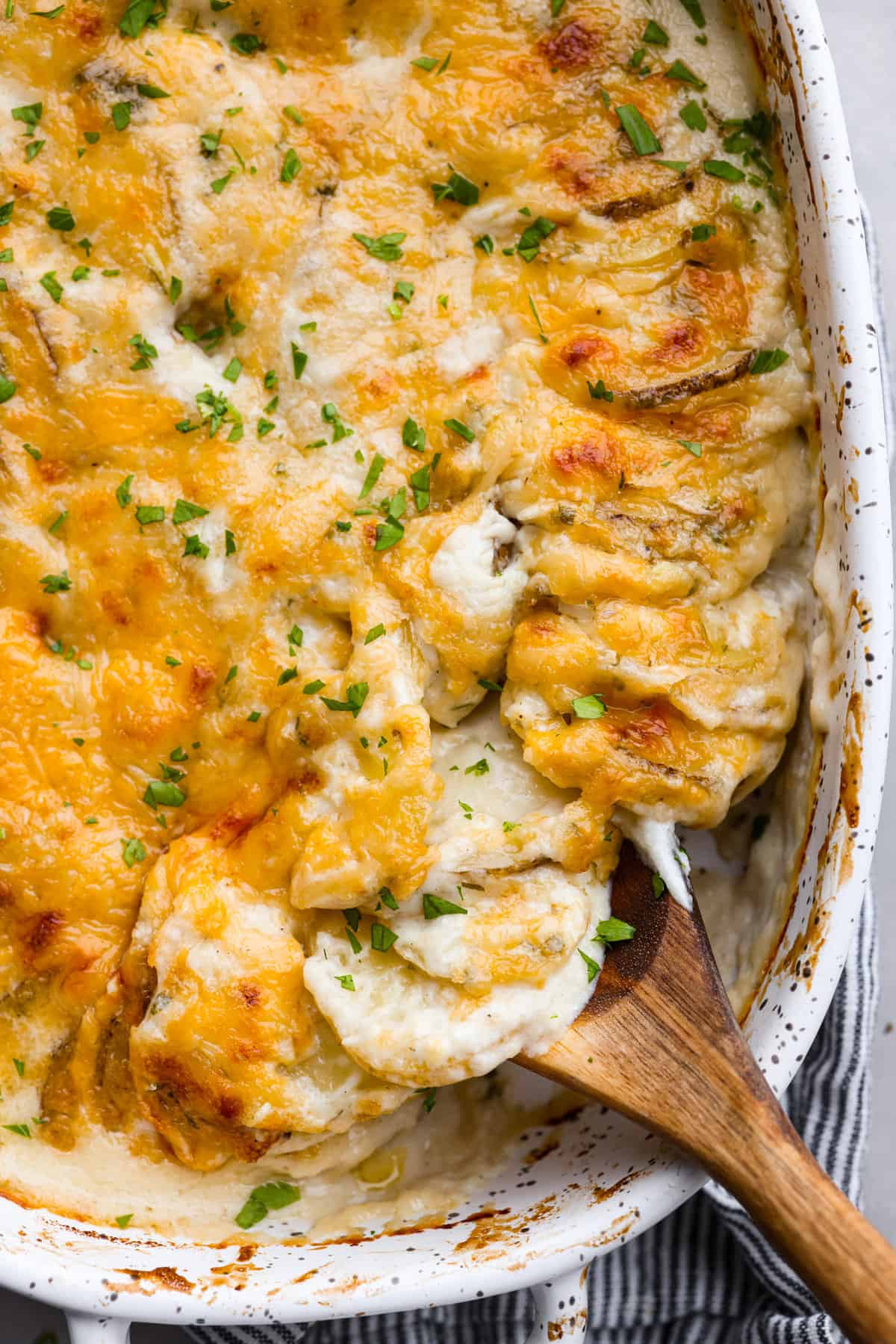 Creamy and cheesy au gratin potatoes are the perfect side dish to any comforting meal. Tender sliced potatoes baked in a rich and flavorful sauce with a golden layer of melty cheese on top. No one will be able to resist these heavenly potatoes. Close up view of au gratin potatoes in a baking dish with a wooden spoon lifting up potatoes.