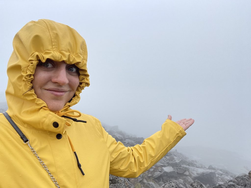 Kate standing in a hooded yellow raincoat, holding her hand out toward a foggy landscape where you can see almost nothing behind her.