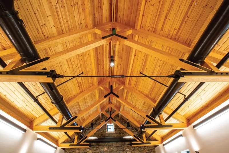View of a rustic wooden ceiling featuring exposed beams and black metal piping, illuminated by pendant lighting.
