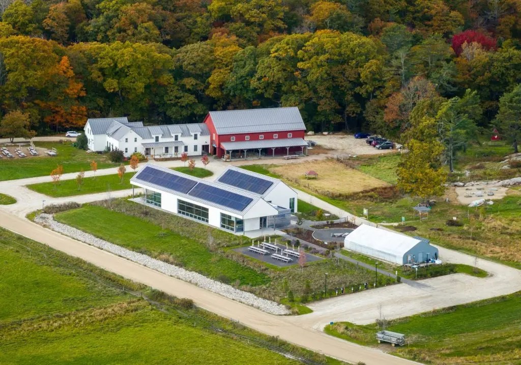 Aerial view of a rural educational farm with solar panels, greenhouses, and a red barn surrounded by vibrant autumn trees, conveying sustainability.