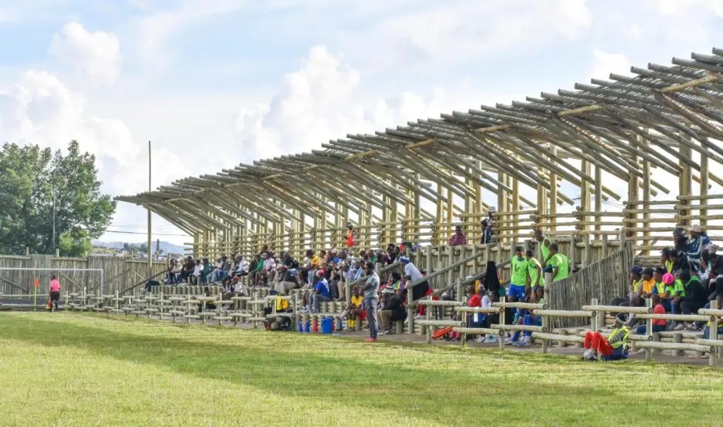 A crowd seated on wooden bleachers under a structured canopy watches an outdoor event on a sunny day, conveying a lively and communal atmosphere.