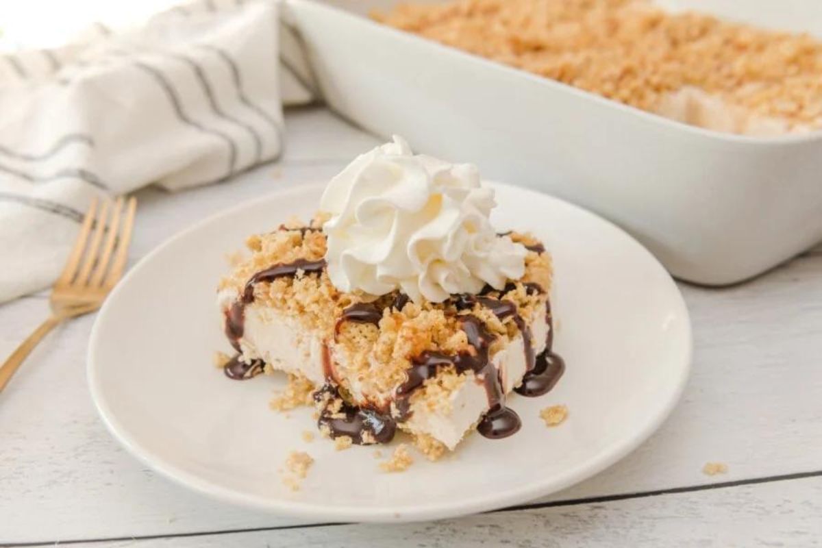 A slice of crumbly dessert topped with whipped cream, ice cream, and drizzled with chocolate sauce, served on a white plate.