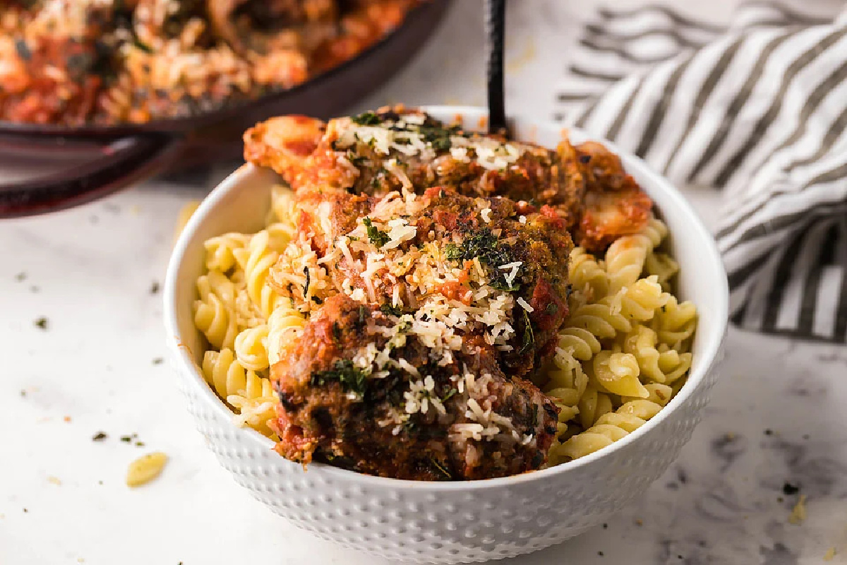 A bowl of pasta topped with tomato sauce, chicken, and sprinkled with herbs and grated cheese, on a marble countertop with a skillet in the background.