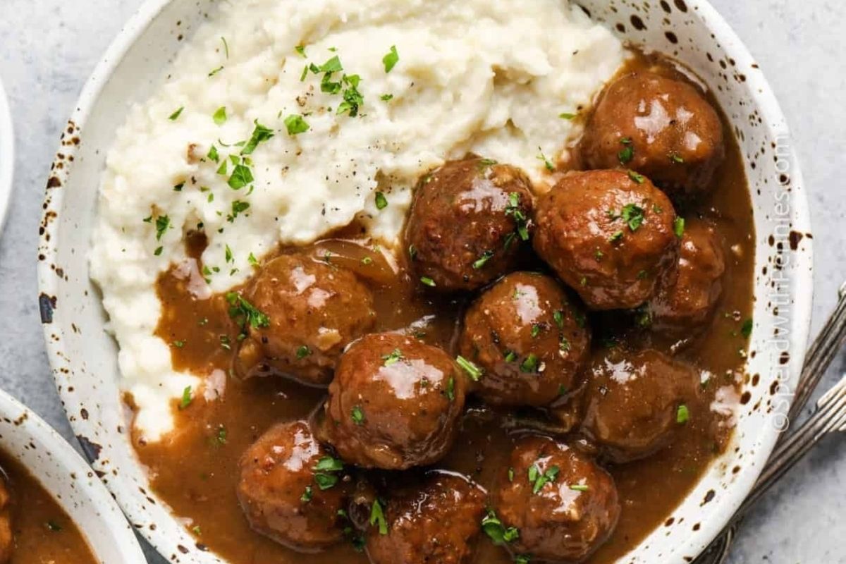 A bowl of meatballs in brown gravy served with mashed potatoes, garnished with chopped parsley - a perfect after-church crockpot recipe.