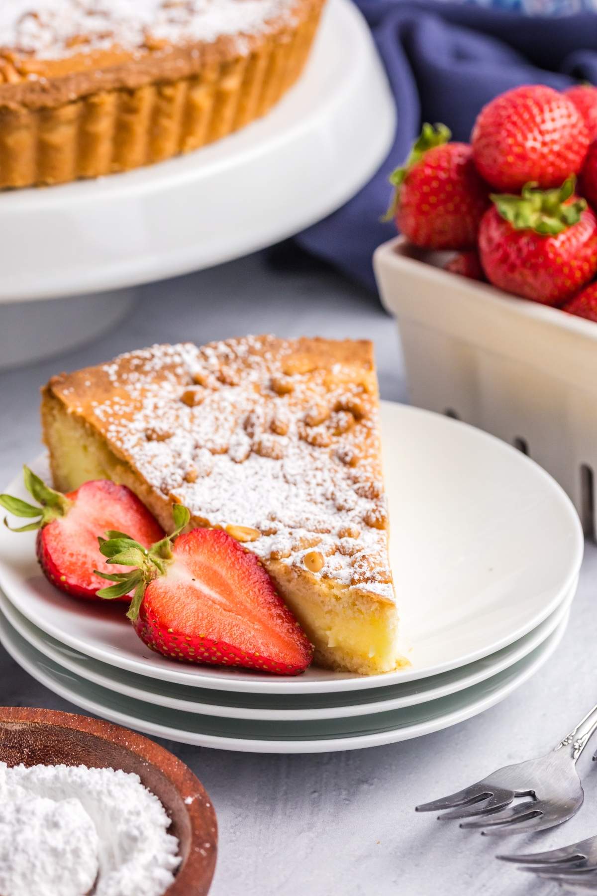 A slice of powdered sugar-dusted torta della nonna (also known as grandmother's cake) on a white plate with two fresh strawberries, next to a bowl of strawberries and a small dish of powdered sugar, on a light surface.